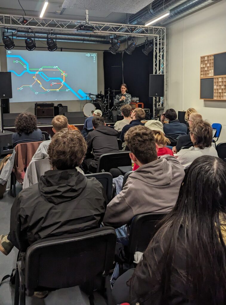 Markus Zierhofer (CEO at Berlin-based game sound and music studio Audio Creatures) delivers a masterclass at BIMM Berlin. In the foreground, a group of students are sitting listening. In the background, you can see Markus who has a video game presented on the board.
